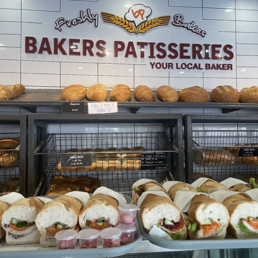 The counter at Bakers Patisseries: the maroon sign on white tiles, loaves along the shelf and a tray of pork rolls at the front